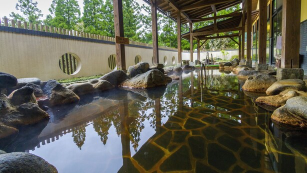 A traditional Japanese onsen with a wooden pavilion, stone bath, and a stone wall with circular cutouts. - Aso Resort Grandvrio Hotel (Aso)