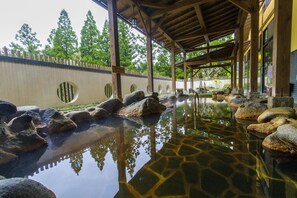 A traditional Japanese onsen with a wooden pavilion, stone bath, and a stone wall with circular cutouts.