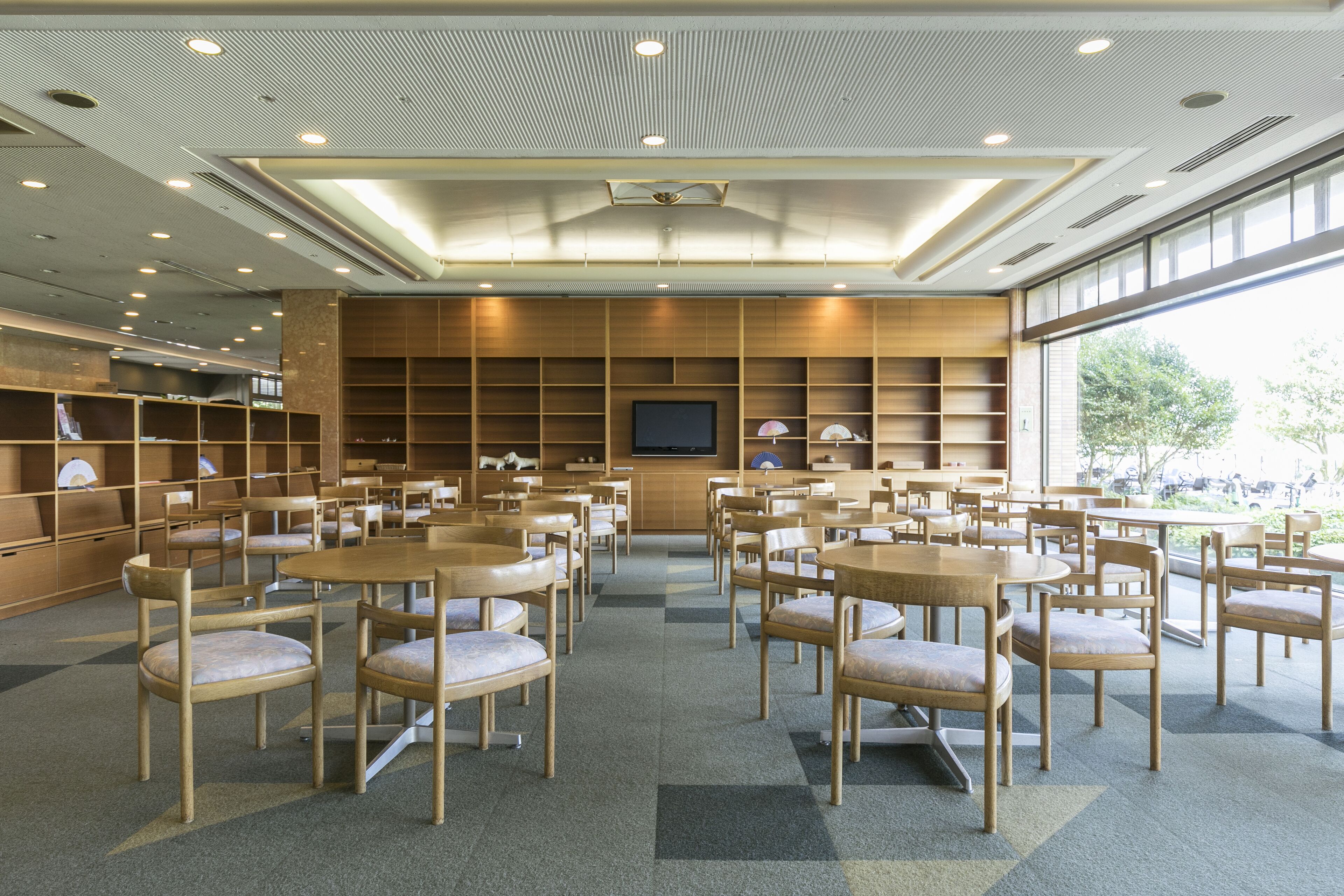 A spacious library with multiple wooden tables and chairs, a large television screen, and bookshelves.