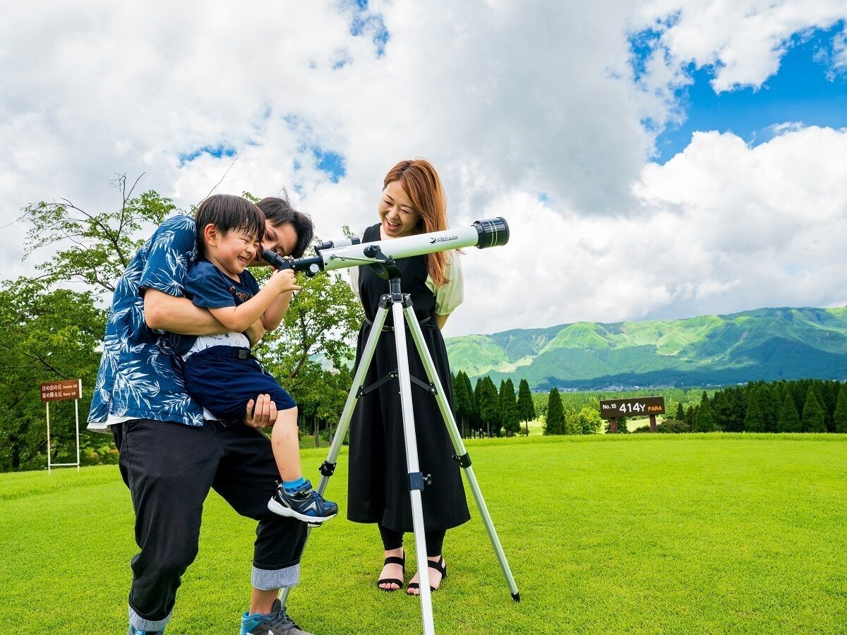 A person using a telescope on a grassy field with mountains in the background.