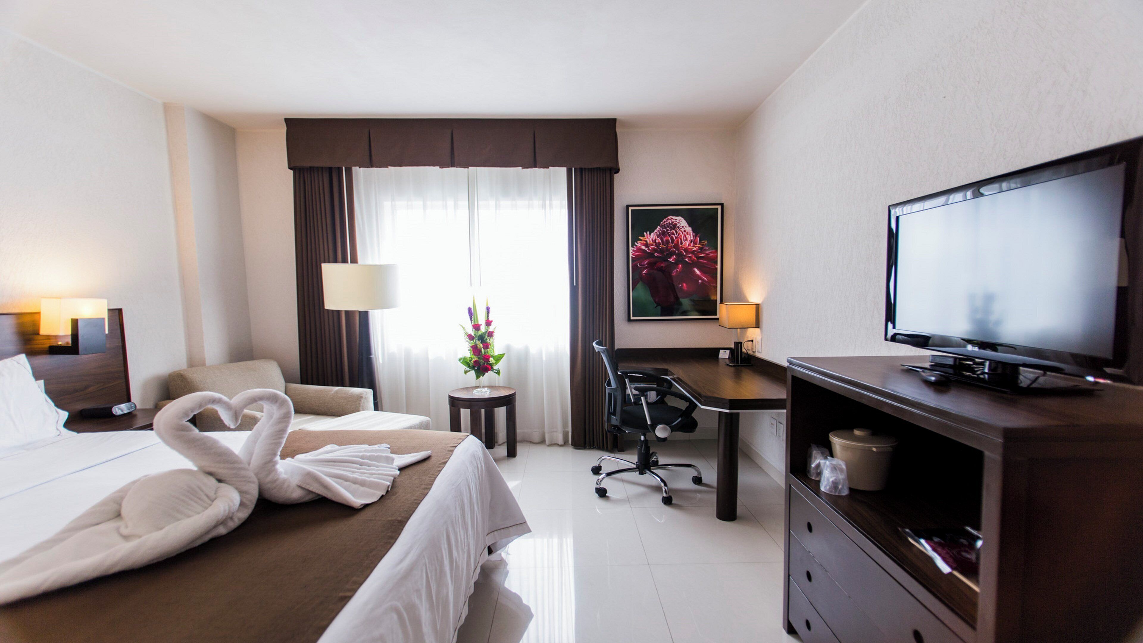 A hotel room with a bed, a desk, a chair, a television, and a painting on the wall.