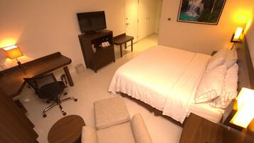 A hotel room with a bed, a desk with a chair, a television, a small table, and a painting on the wall.