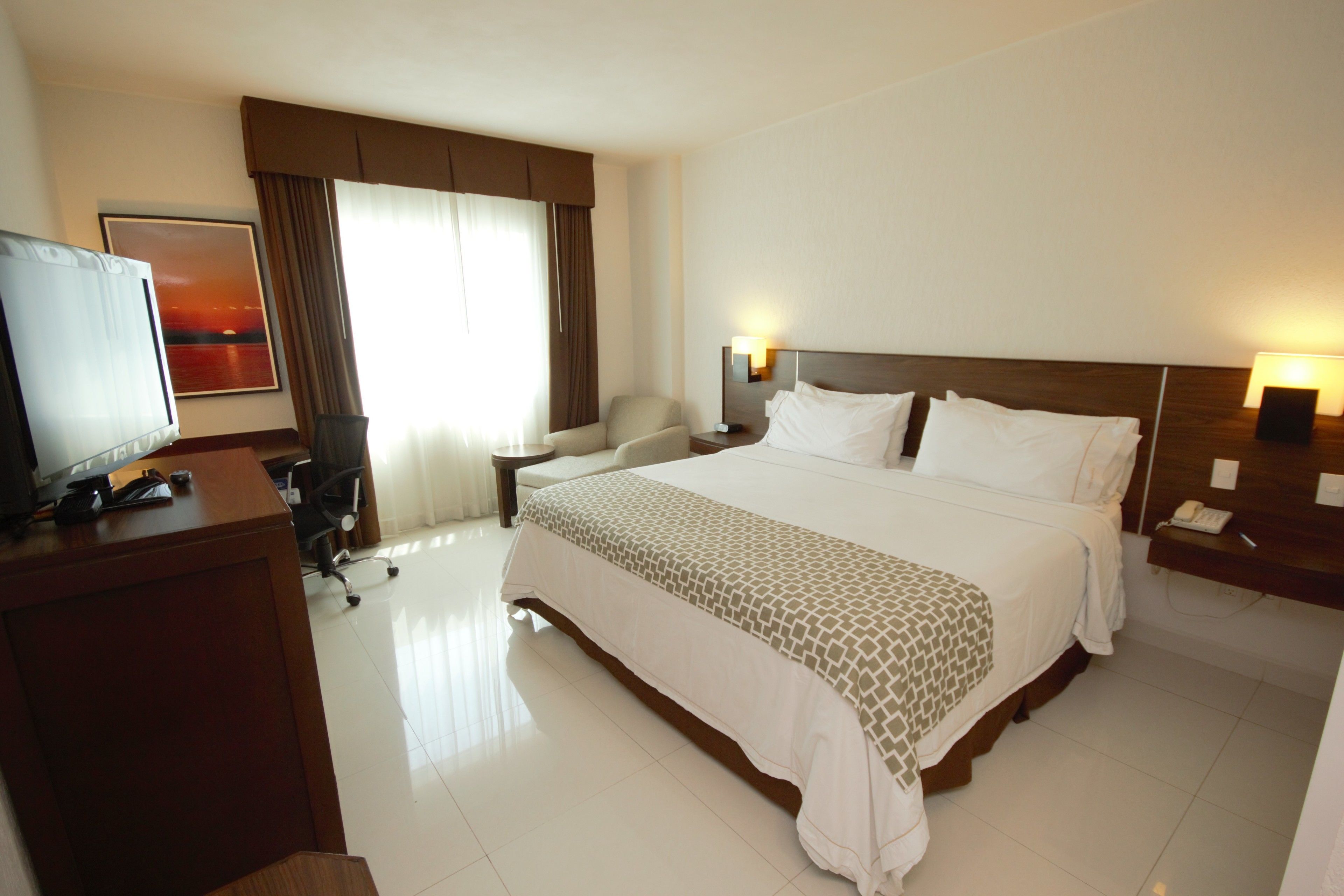 A hotel room with a large bed, a desk with a television, a chair, and a nightstand with a lamp.