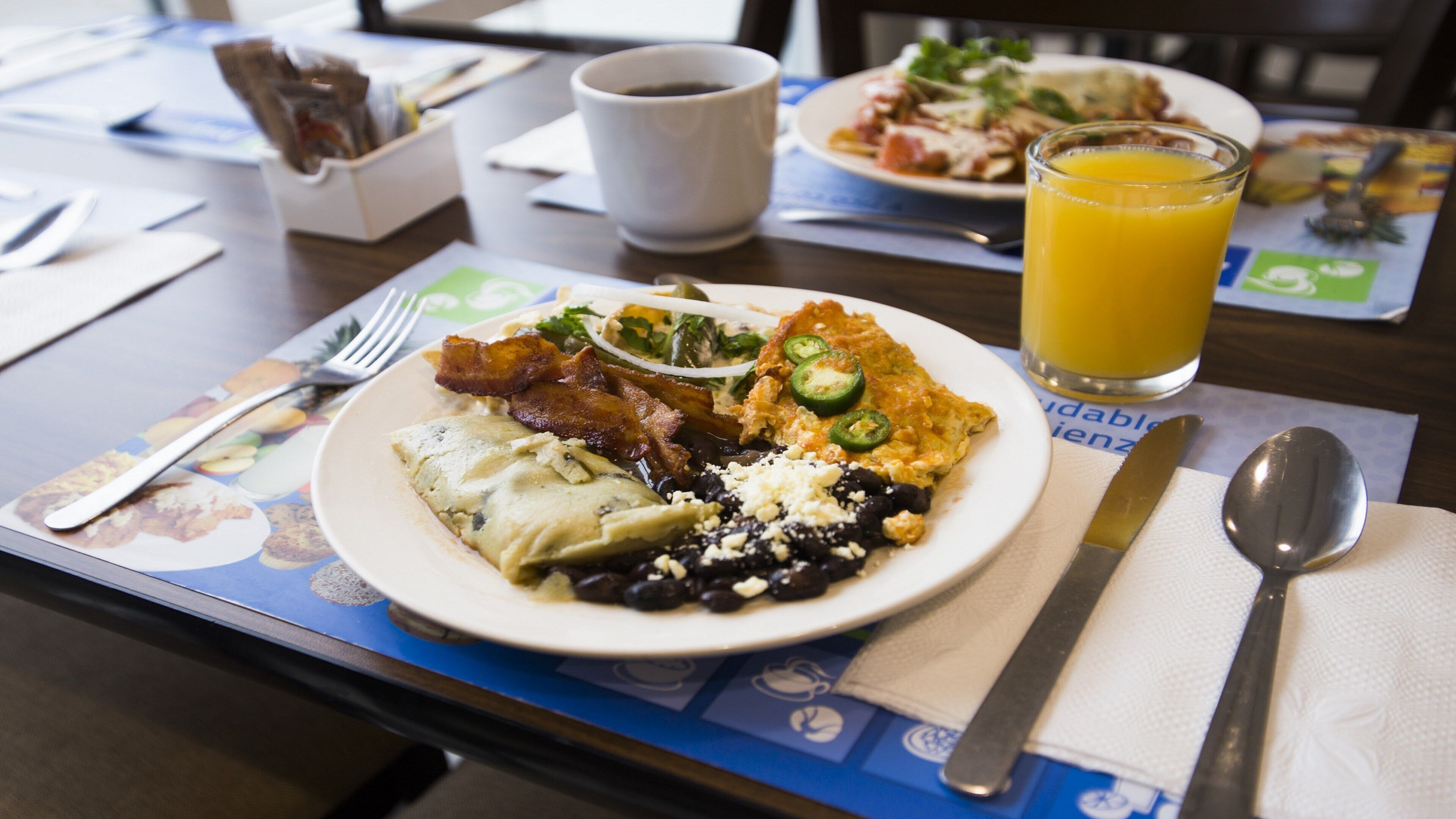 Un plato de desayuno con huevos, tocino, frijoles negros y un vaso de jugo de naranja sobre una mesa con servilleta, tenedor y cuchara.