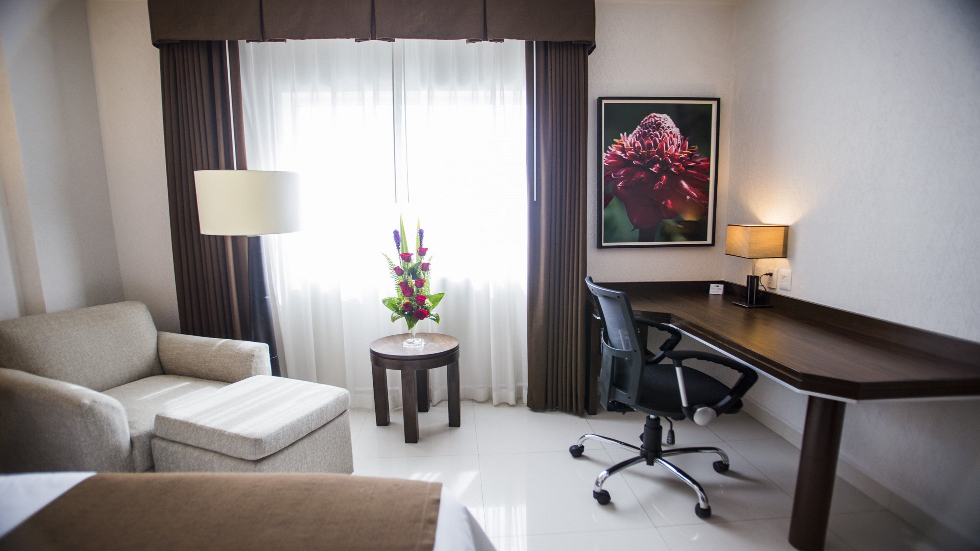 A hotel room with a sofa, a desk with a chair, a small table with a flower arrangement, and a painting on the wall.