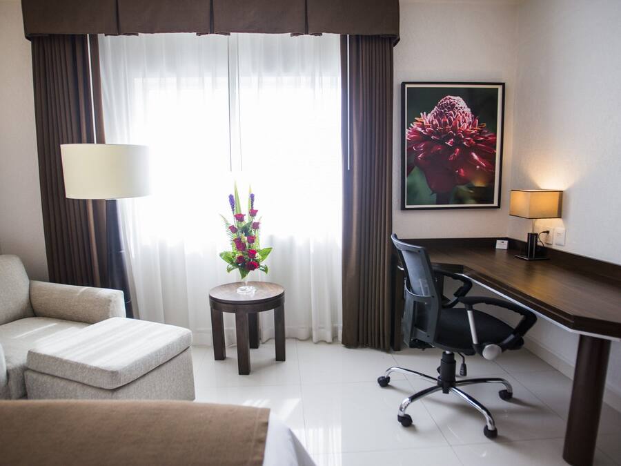A hotel room with a sofa, a desk with a chair, a small table with a flower arrangement, and a painting on the wall.