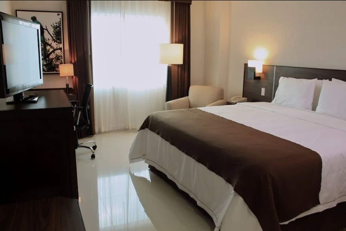 A hotel room with a bed, a desk with a computer, a chair, a lamp, and a window with curtains.