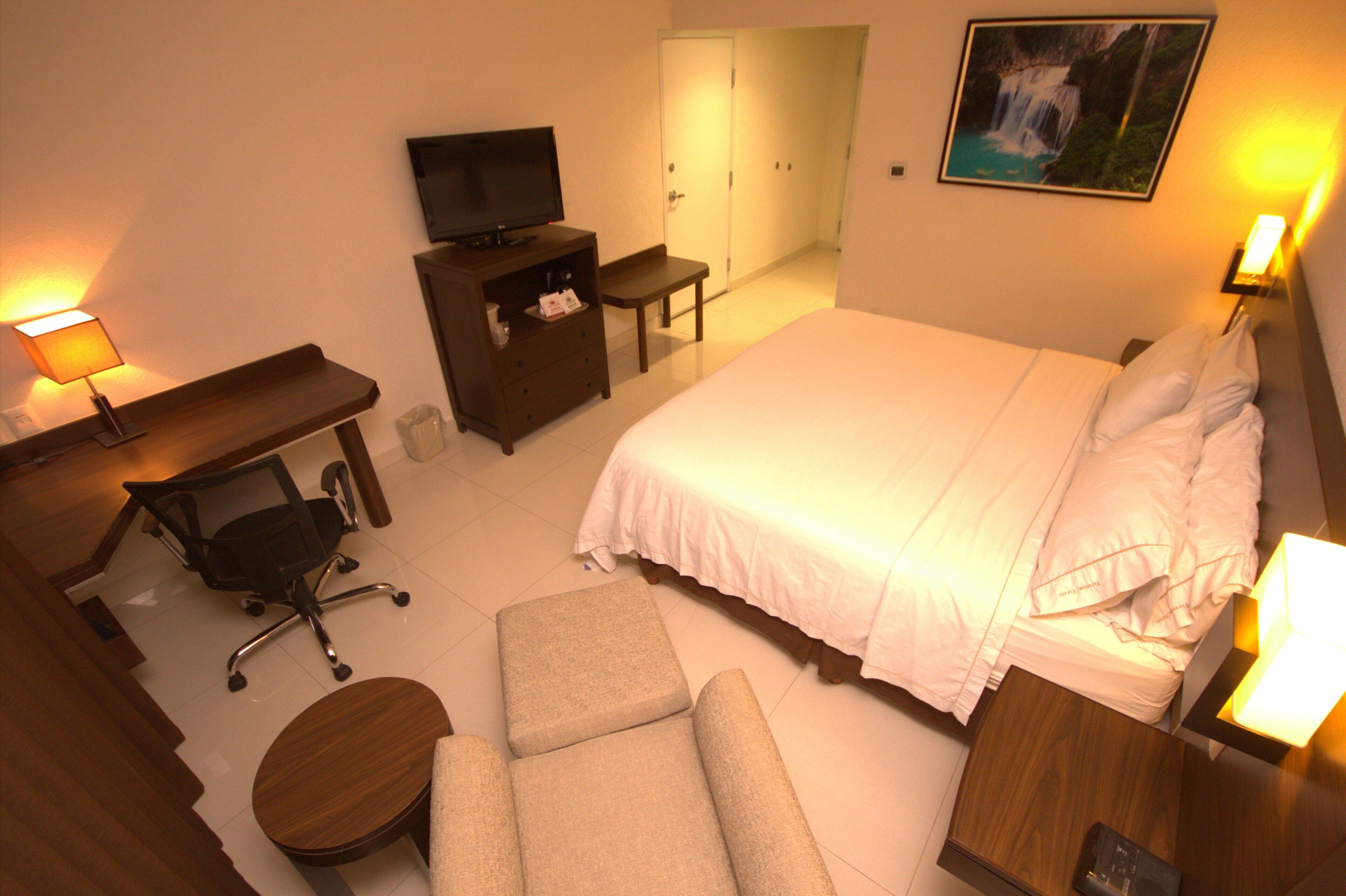 A hotel room with a bed, a desk, a chair, a television, and a painting on the wall.