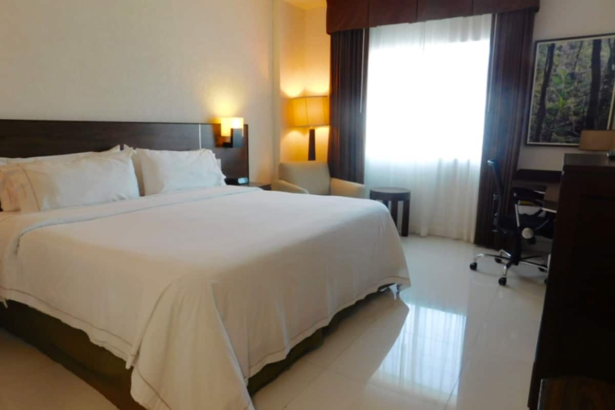 A hotel room with a large bed, two bedside lamps, a desk with a chair, and a window with curtains.