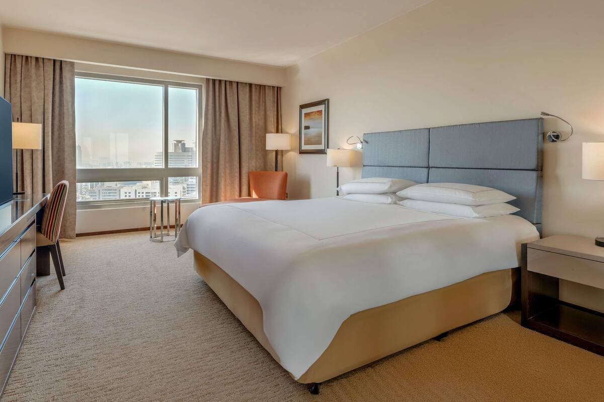 A hotel room with a large bed, a desk, a chair, and a view of the city through the window.