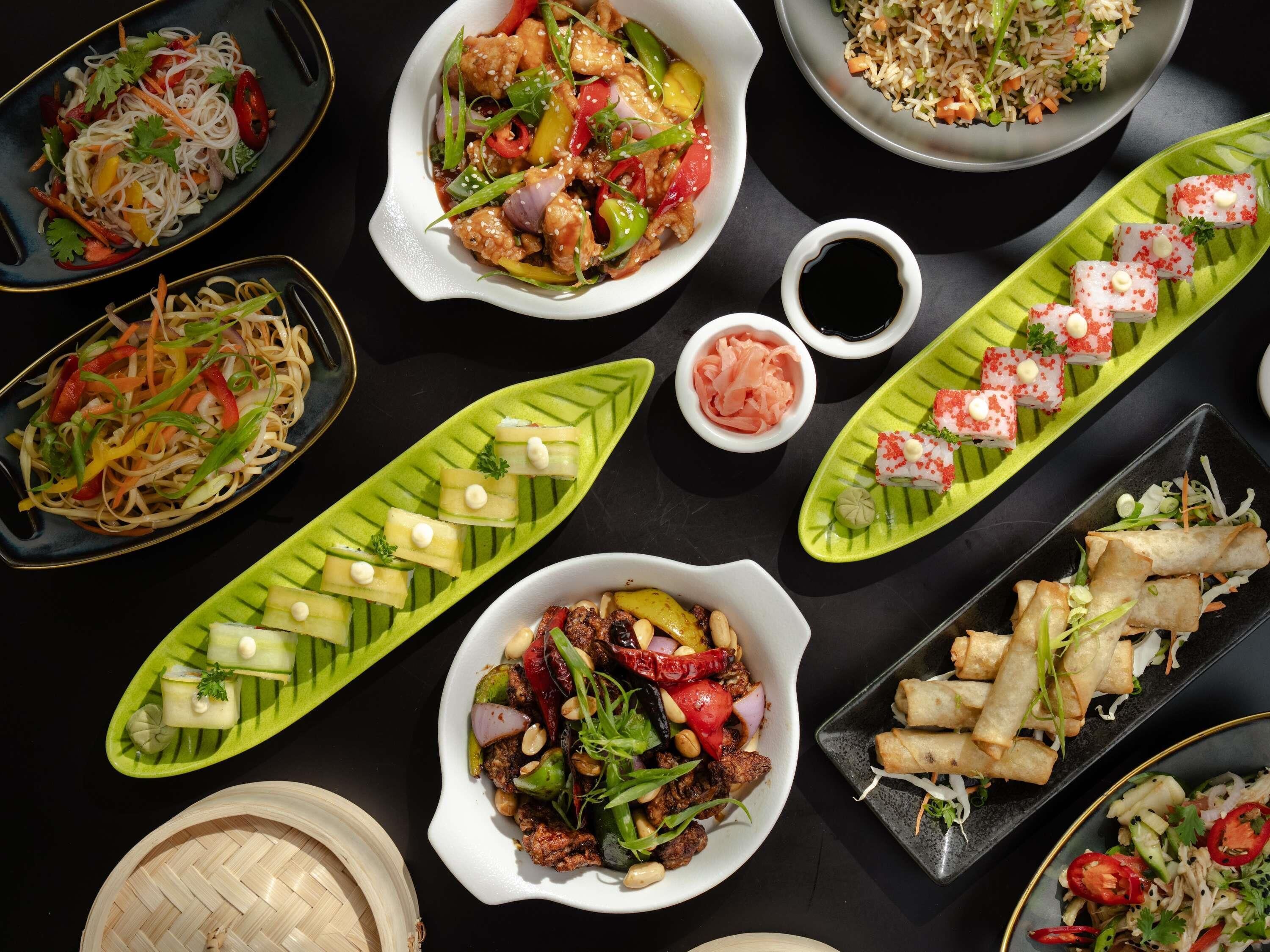 Diverse gerechten, zoals noedels, roerbakgroenten en sushi, op een zwarte tafel.