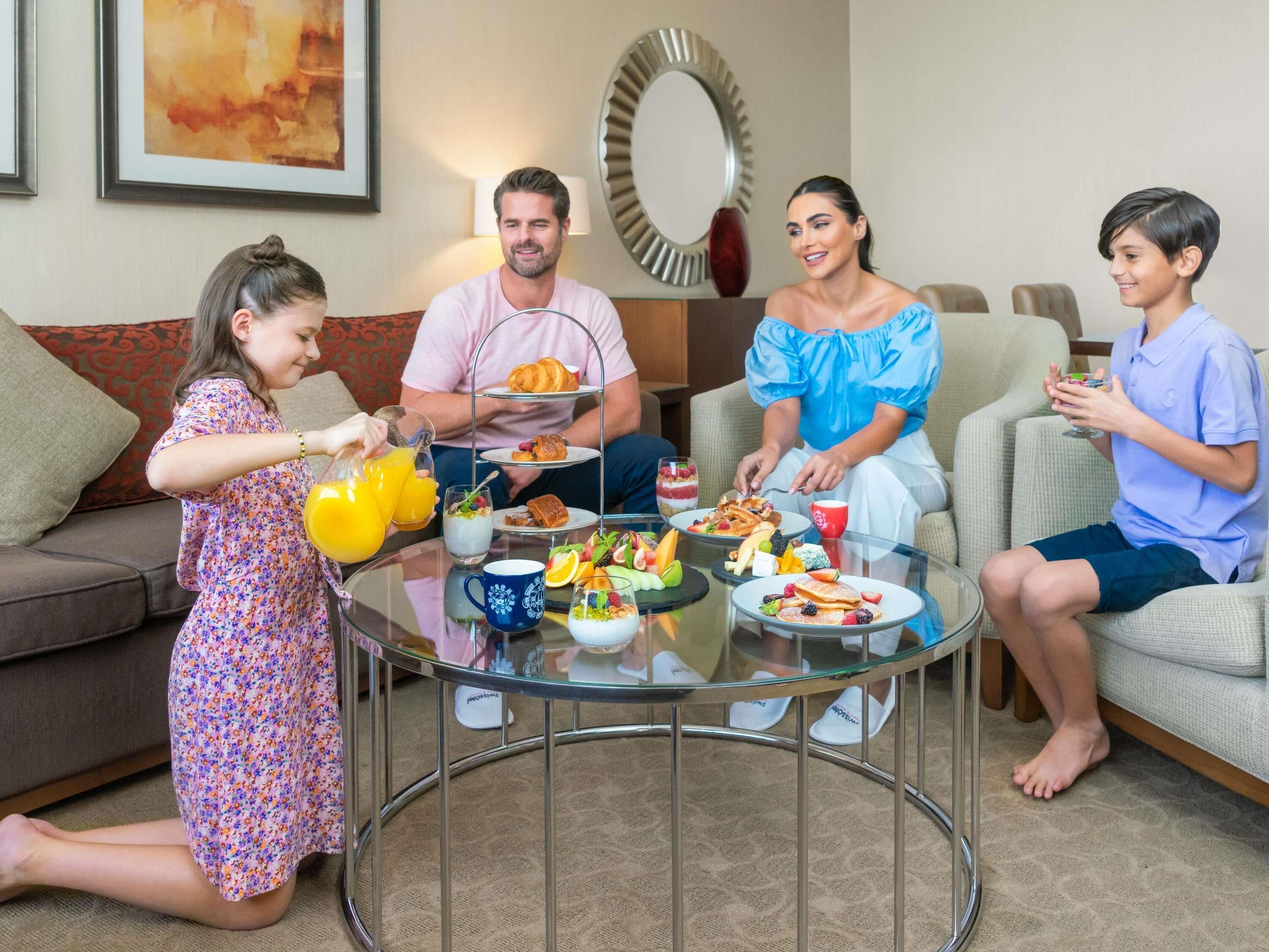 Une famille est assise autour d’une table dont le plateau est en verre, dans un salon où l’on aperçoit un canapé et, au fond, un miroir décoratif.