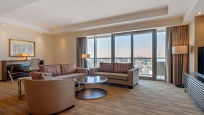 A modern hotel room with a large window, a sofa set, a coffee table, and a desk with a lamp.