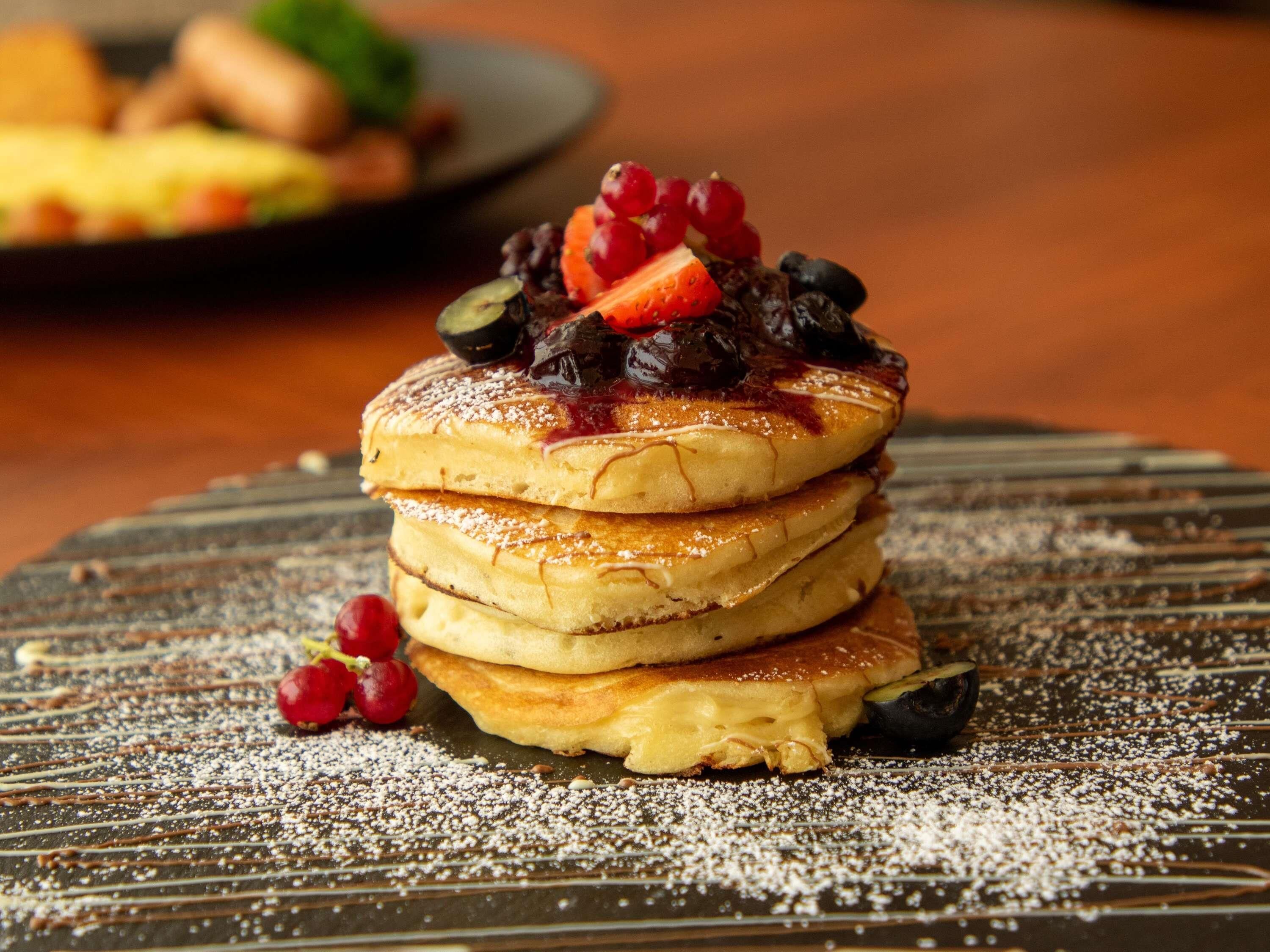 A stack of pancakes with fruit toppings and powdered sugar, served on a dark plate.