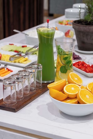 Una mesa de buffet con diversas frutas, un batido verde y vasos vacíos sobre una encimera de mármol.