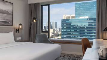 A hotel room with a bed, a desk, a chair, a window with a city view, and a lamp.