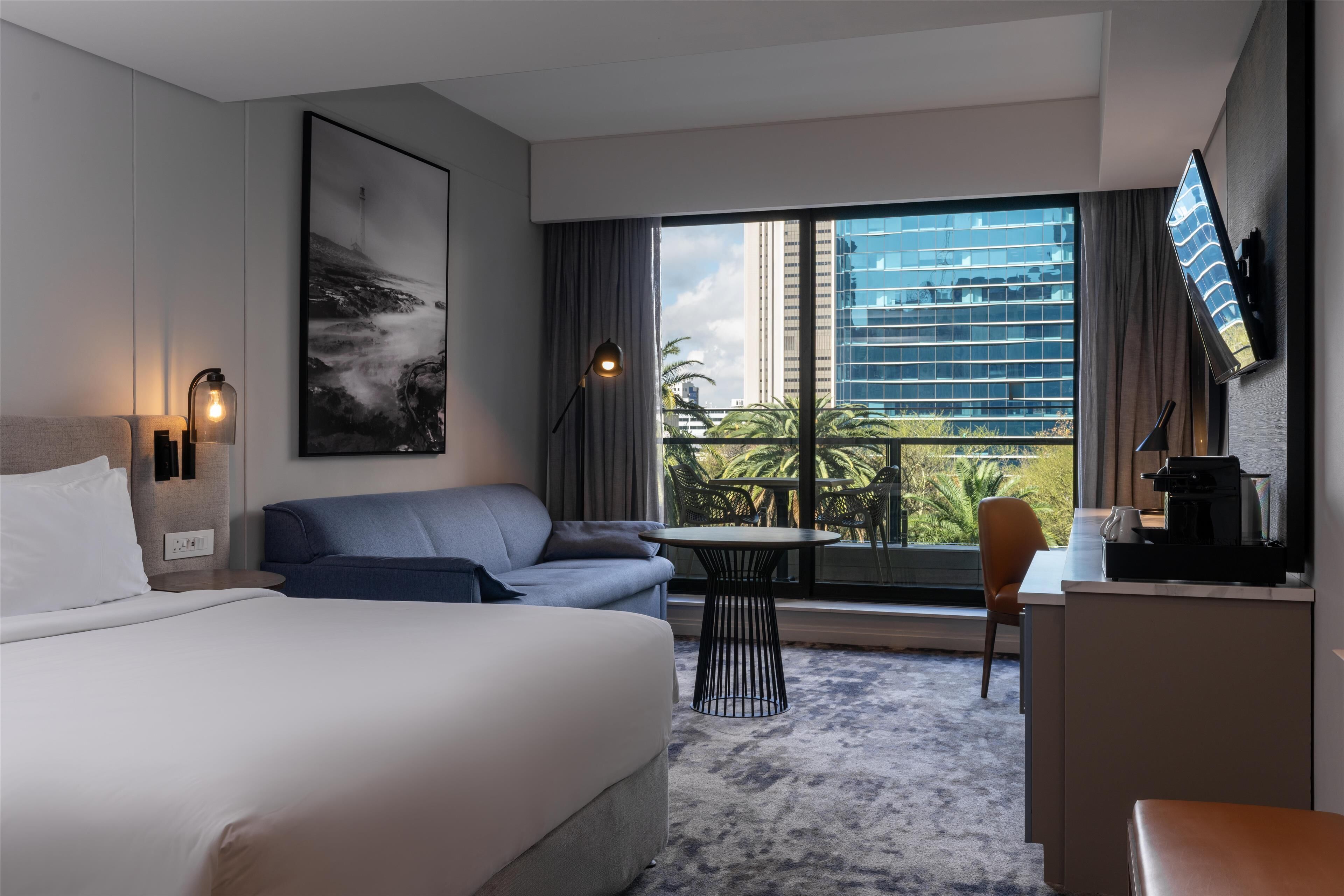 Habitación de hotel con cama, sofá, escritorio, silla, mesa y un ventanal con vistas a edificios y zonas verdes.