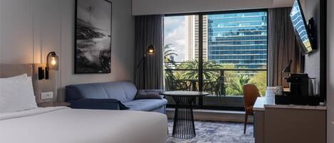 Habitación de hotel con cama, sofá, escritorio, silla, mesa y un ventanal con vistas a edificios y zonas verdes.