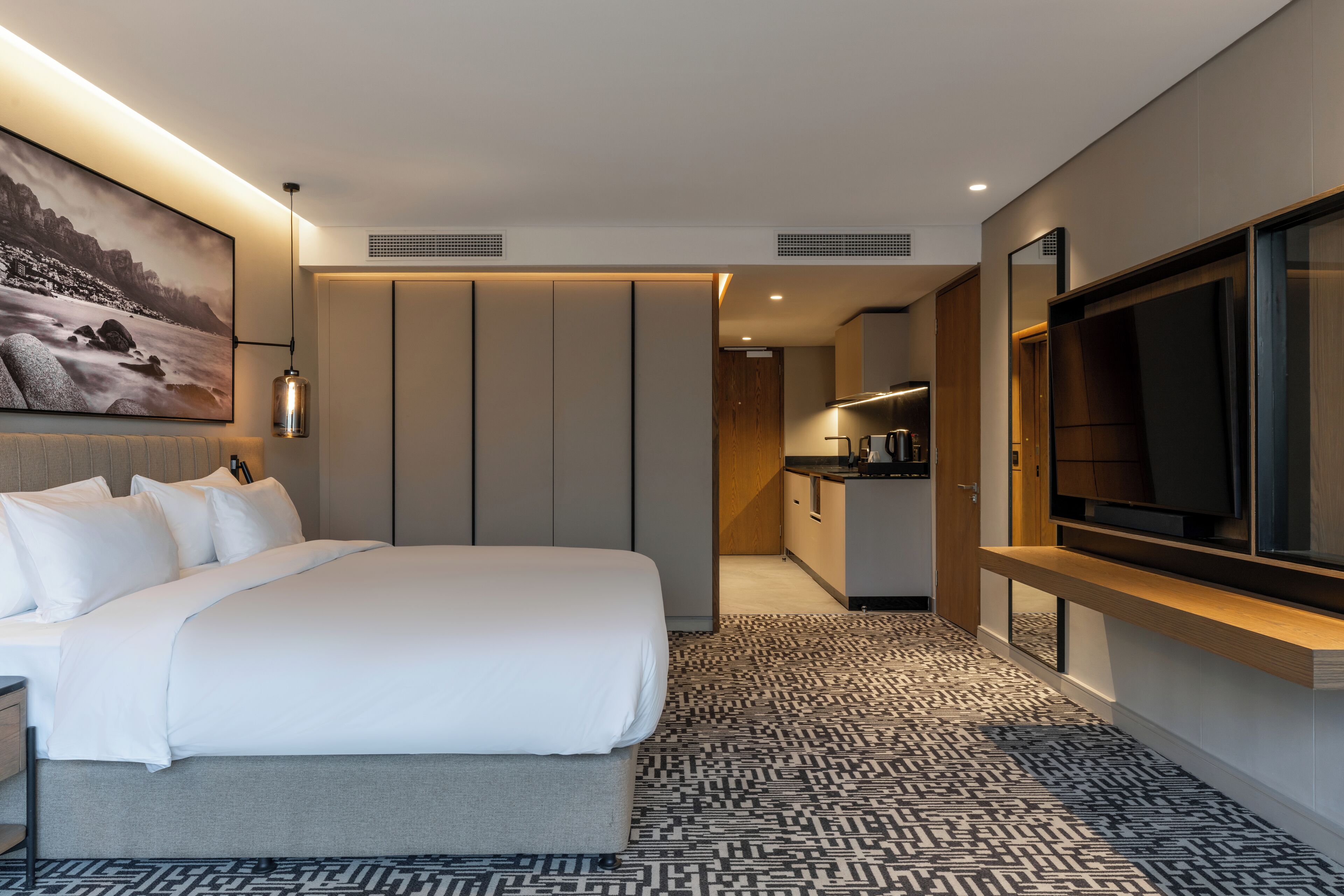A hotel room with a large bed, a TV, a painting, and a kitchenette.