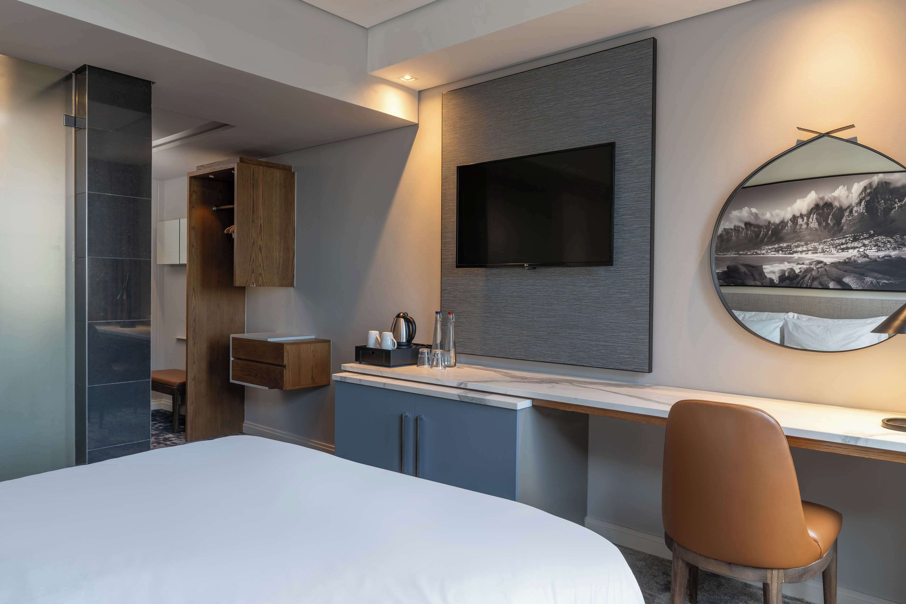A modern hotel room with a bed, a desk, a chair, a wardrobe, a mirror, and a flat-screen TV.