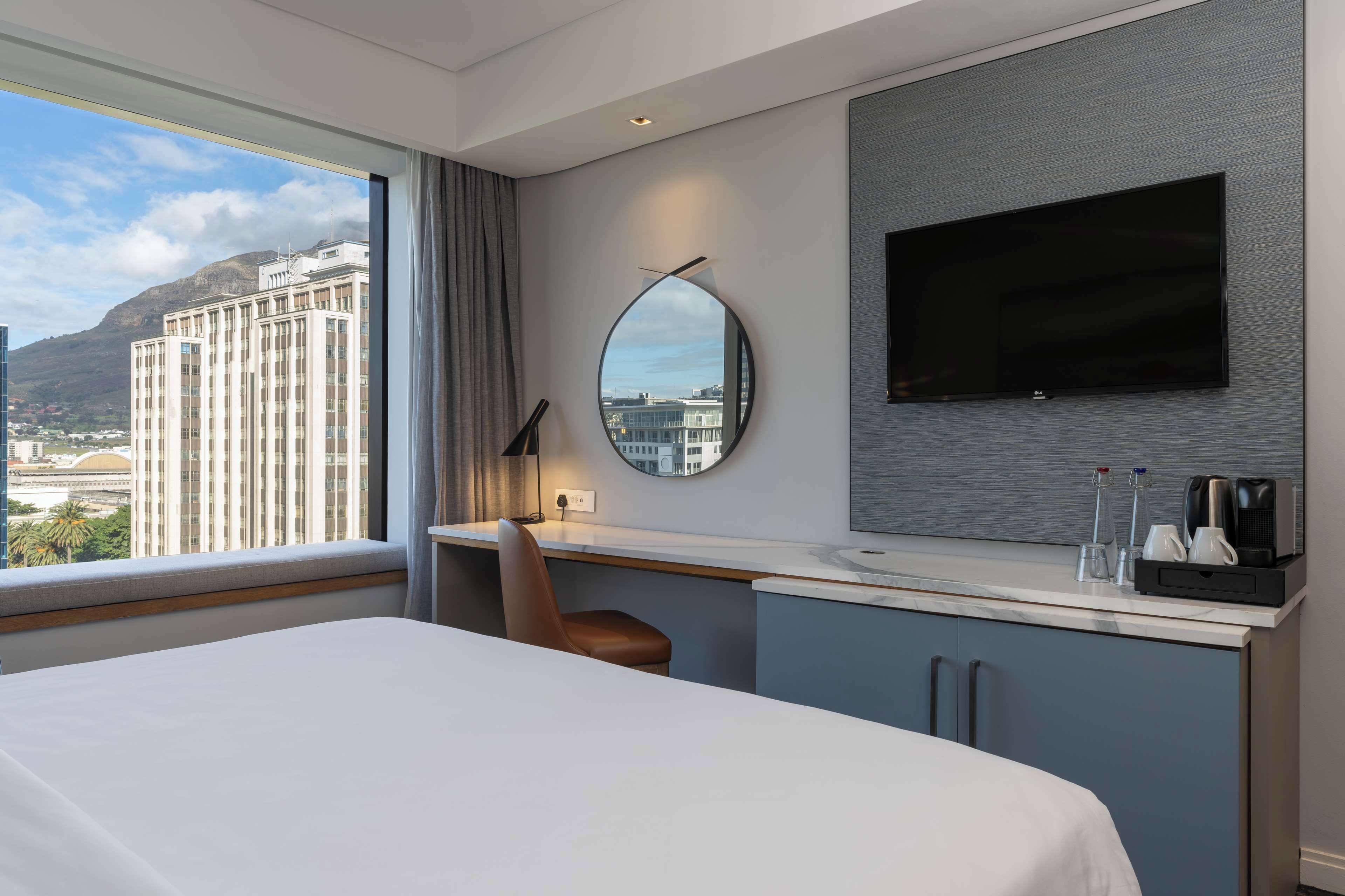 A modern hotel room with a large bed, a desk with a chair, a flat-screen TV, a round mirror, and a view of a cityscape through the window.