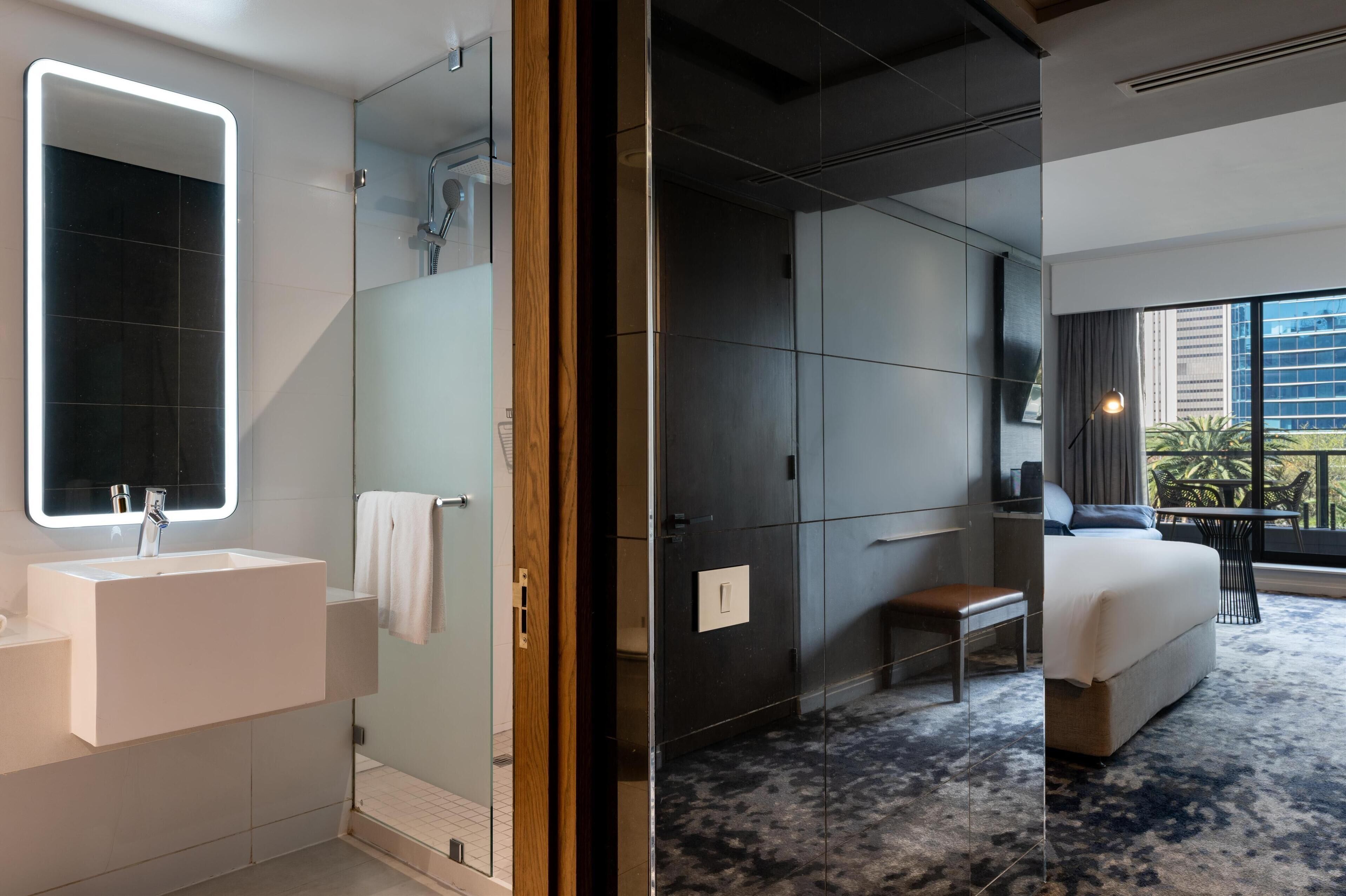 Een moderne hotelkamer met een grote spiegel, een wit wastafelmeubel, een douche met glazen wand en een bed met uitzicht, plus een klein tafeltje.