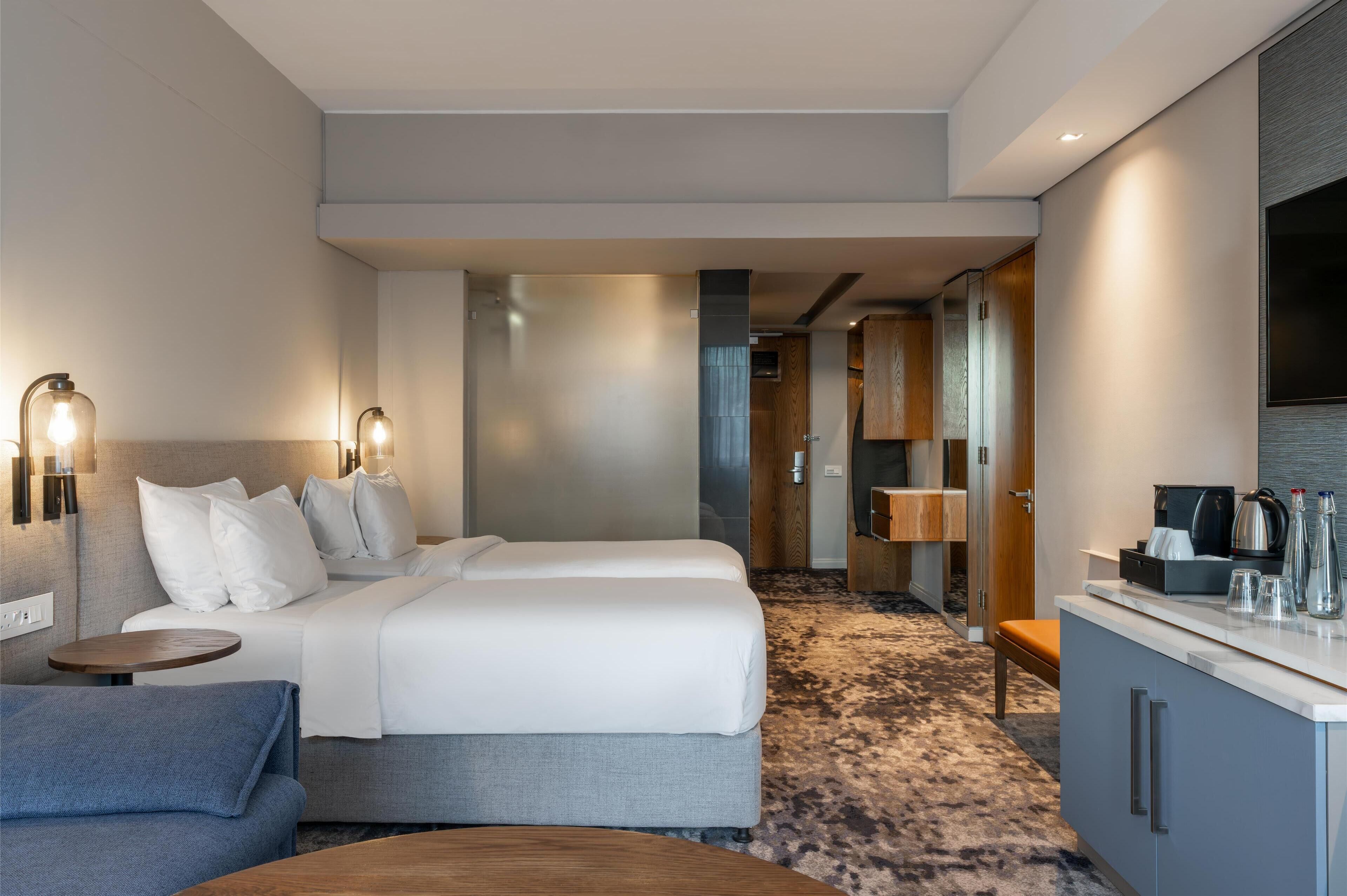 Quarto de hotel com duas camas, área de estar, cozinha compacta e banheiro visível através de uma porta aberta.