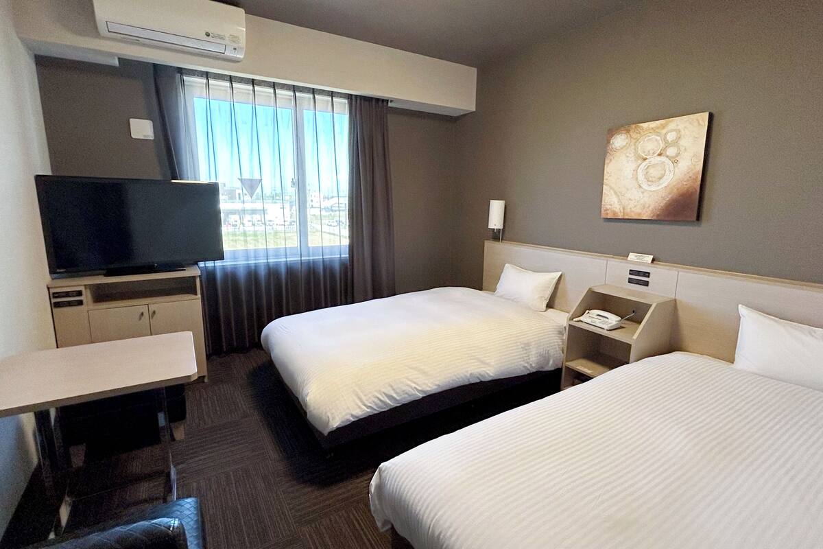 A hotel room with two beds, a TV, a desk, and a window with curtains.