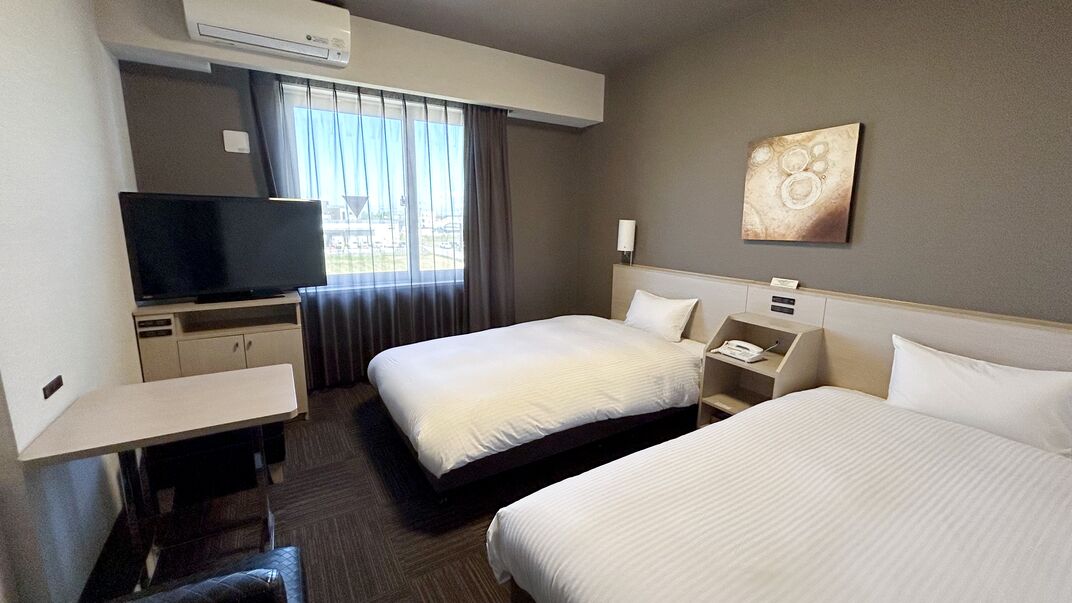 A hotel room with two beds, a TV, a desk, and a window with curtains.