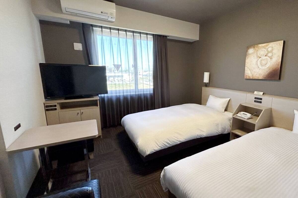 A hotel room with two beds, a TV, a desk, and a window with curtains.