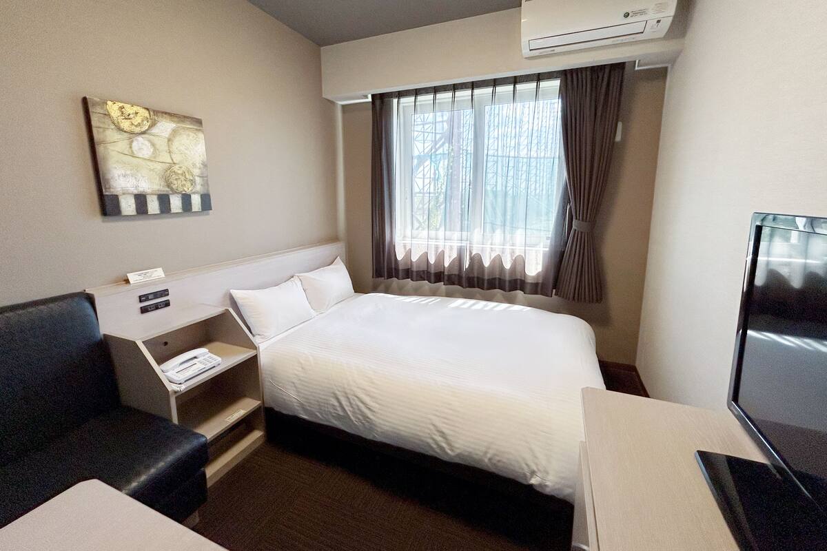 A hotel room with a bed, a desk, a chair, an air conditioner, a window with curtains, and a painting on the wall.