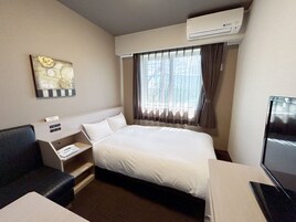 Habitación de hotel con cama, escritorio, silla, aire acondicionado, ventana con cortinas y un cuadro en la pared.