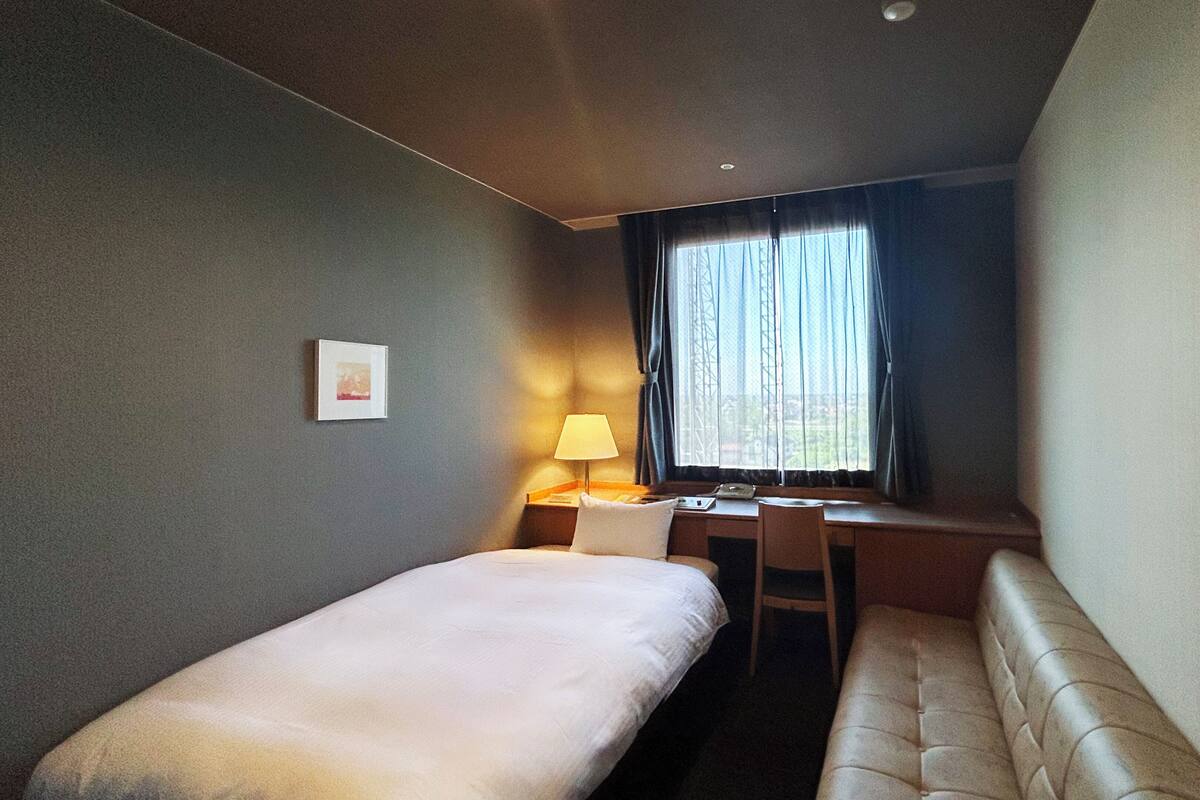 A hotel room with a single bed, a desk, a chair, and a window with curtains.