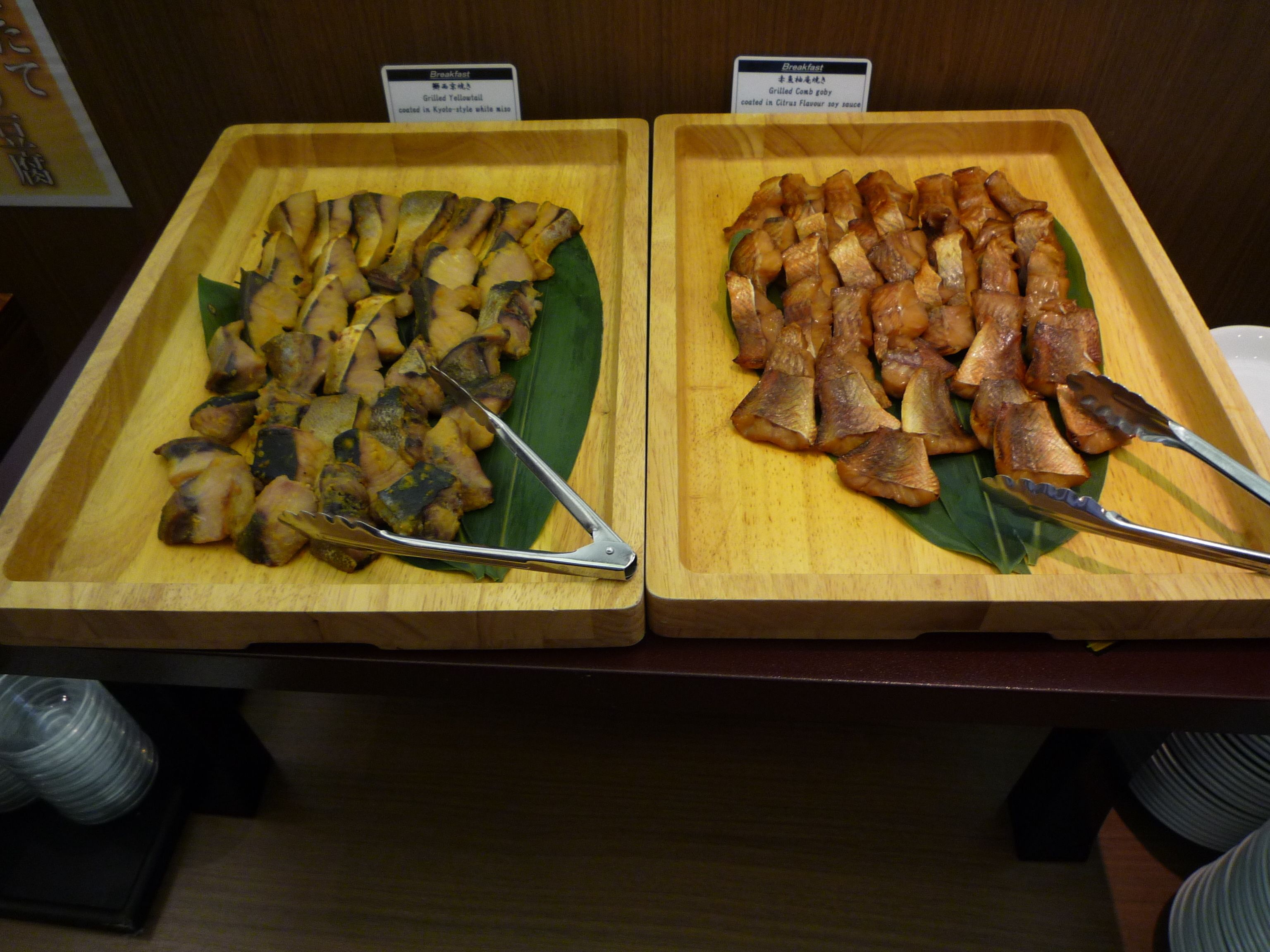 Two wooden trays of food with tongs, labeled as 'Grilled Yellowtail' and 'Grilled Lamb'.