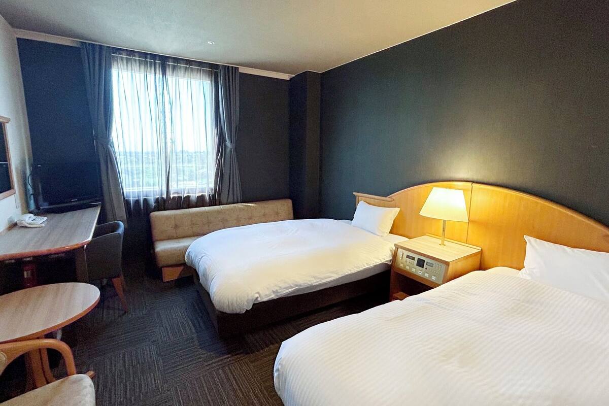A hotel room with two beds, a desk, a chair, a TV, and a window with curtains.