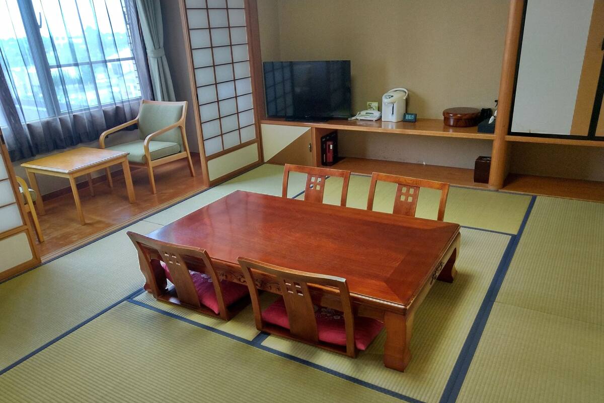 A traditional Japanese room with tatami flooring, a wooden table with four chairs, a television, and a shelf with various items.