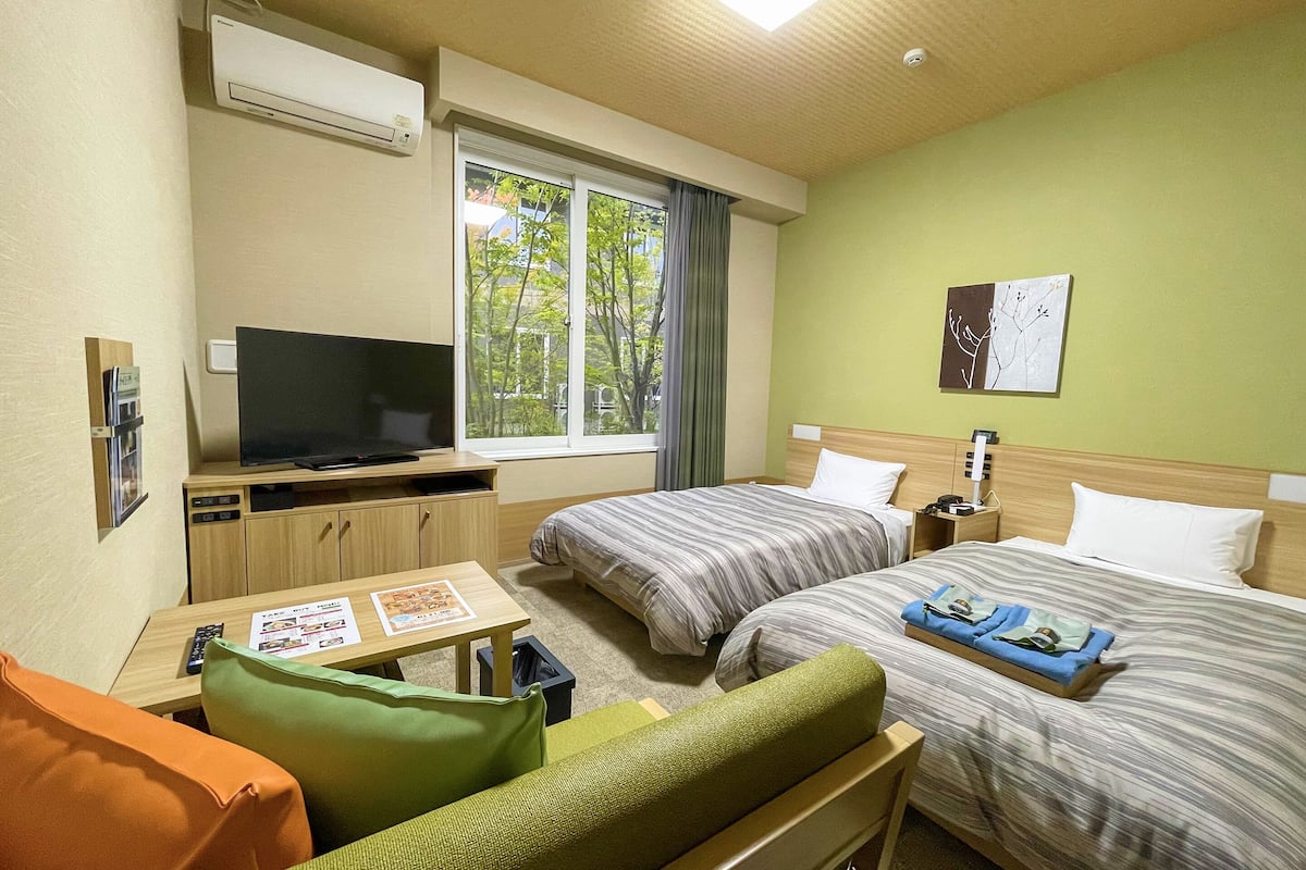 A hotel room with two beds, a TV, a small table, and a window with a view of trees.