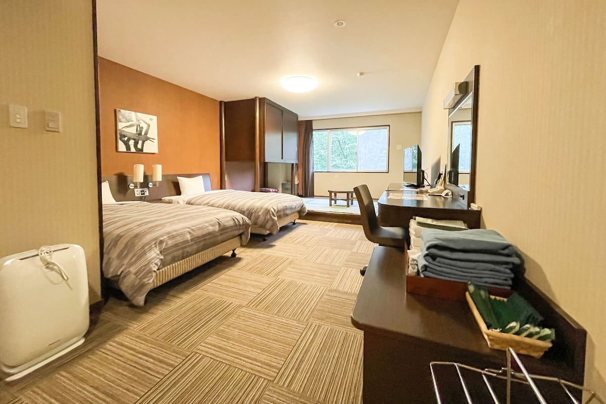 A hotel room with two beds, a desk, a chair, a small table, a mirror, and a window.