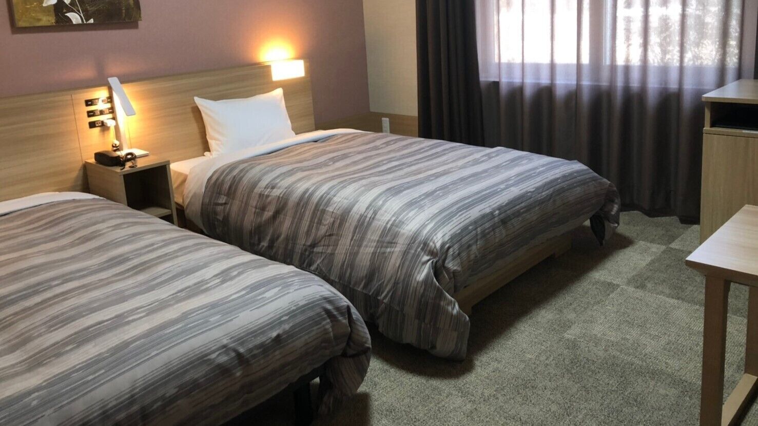 Una camera d'albergo con due letti, un comodino con una lampada, una scrivania e una finestra con tende.