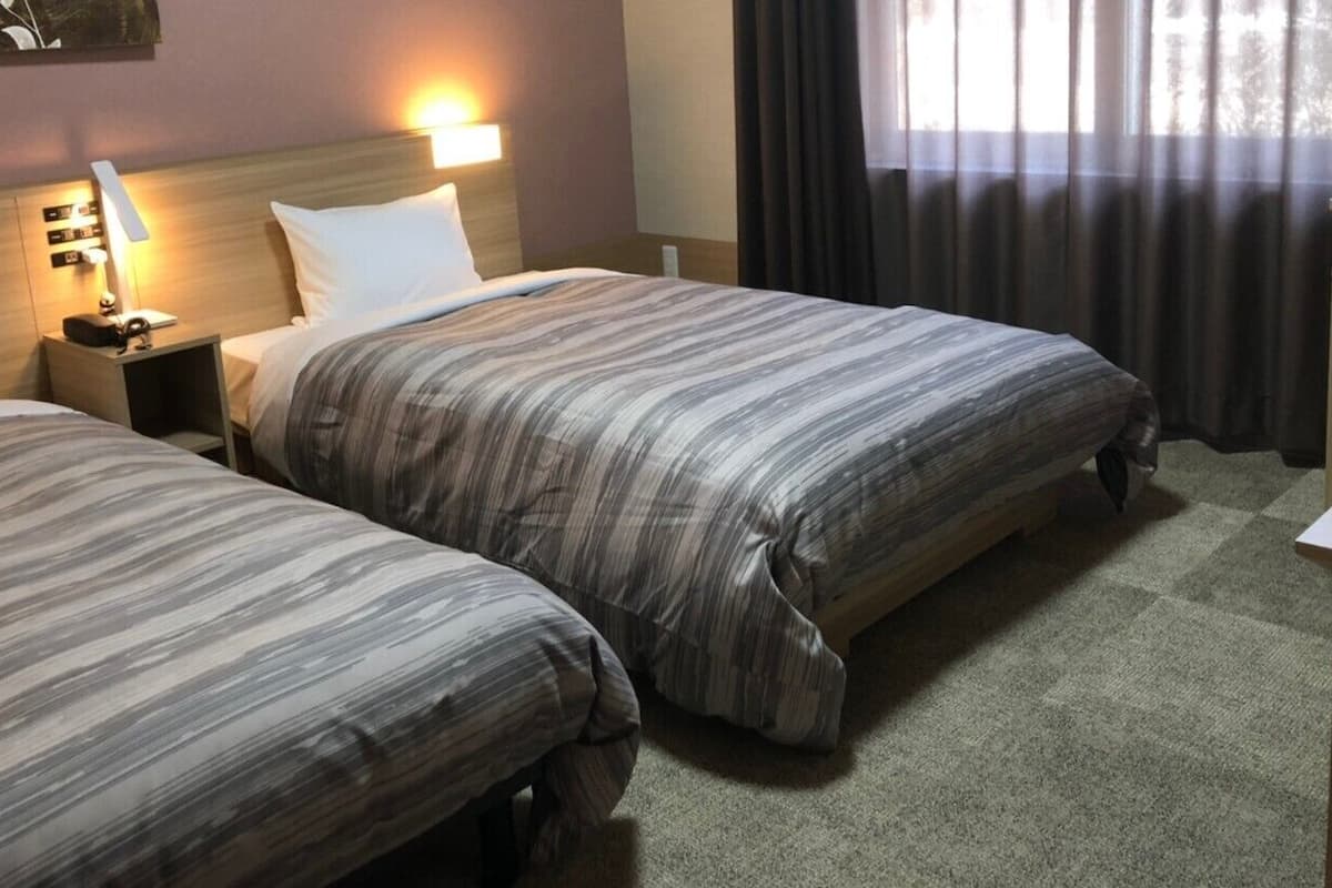 A hotel room with two beds, a nightstand with a lamp, a desk, and a window with curtains.
