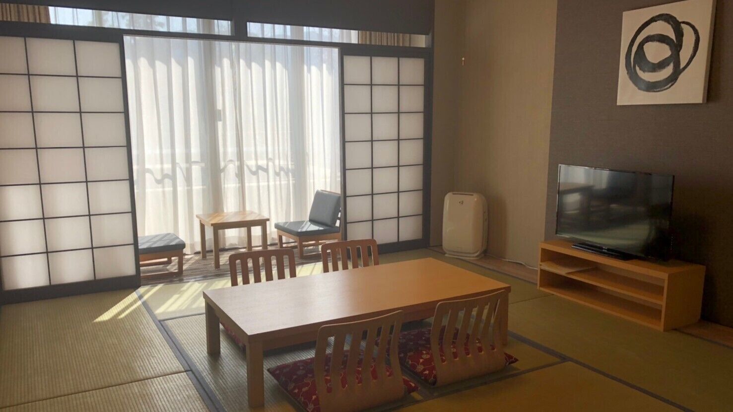 Una tradizionale stanza in stile giapponese con pavimento in tatami, un tavolino con sedie, un televisore e una porta scorrevole che dà su un panorama esterno.