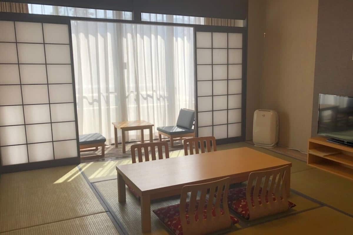 A traditional Japanese-style room with tatami flooring, a low table with chairs, a television, and a sliding door with a view to the outside.