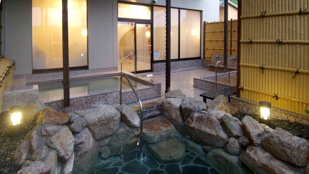 A stone hot tub surrounded by rocks, with a wooden deck and steps leading to a building with large windows. - Route-Inn Grantia Dazaifu (Dazaifu)