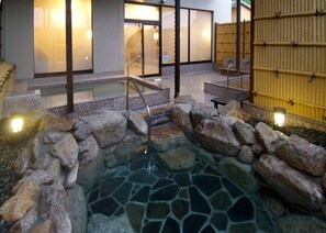 A stone hot tub surrounded by rocks, with a wooden deck and steps leading to a building with large windows. - Route-Inn Grantia Dazaifu (Dazaifu)