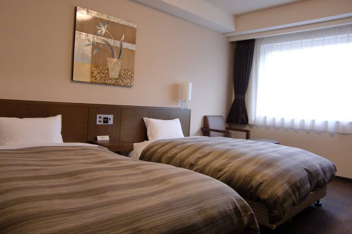 Hotellihuone, jossa on kaksi sänkyä, yöpöytä, lamppu, taulu ja ikkuna, jossa on verhot.
