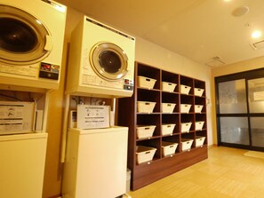 A laundry room with a front-loading washing machine, a dryer, and a shelf with baskets. - Hotel Route-Inn Yokohama Bashamichi (Yokohama)