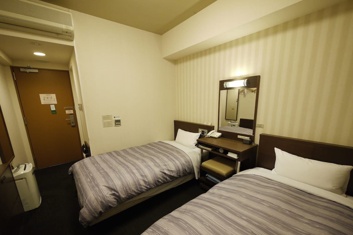 Une chambre d’hôtel avec deux lits, une table de chevet avec un téléphone, un miroir et une porte donnant sur une salle de bain.
