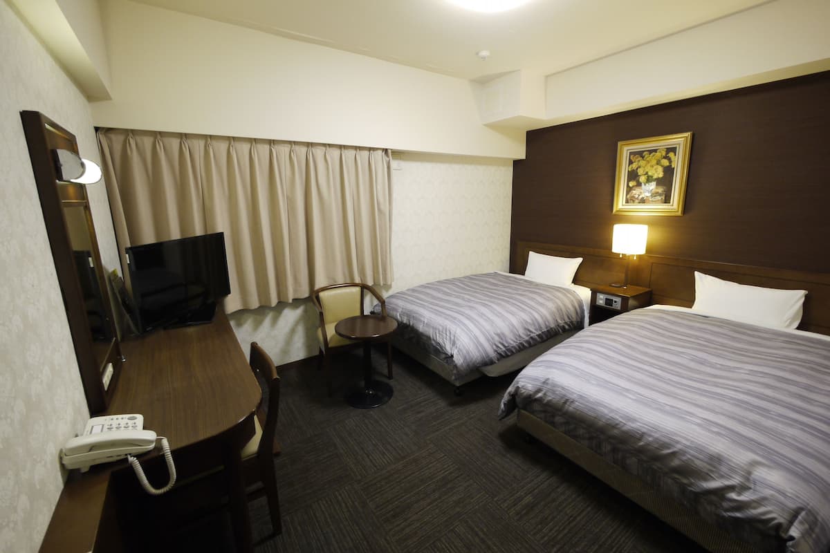 Une chambre d’hôtel avec deux lits, un bureau avec un téléphone, une chaise, une télévision et un tableau au mur.