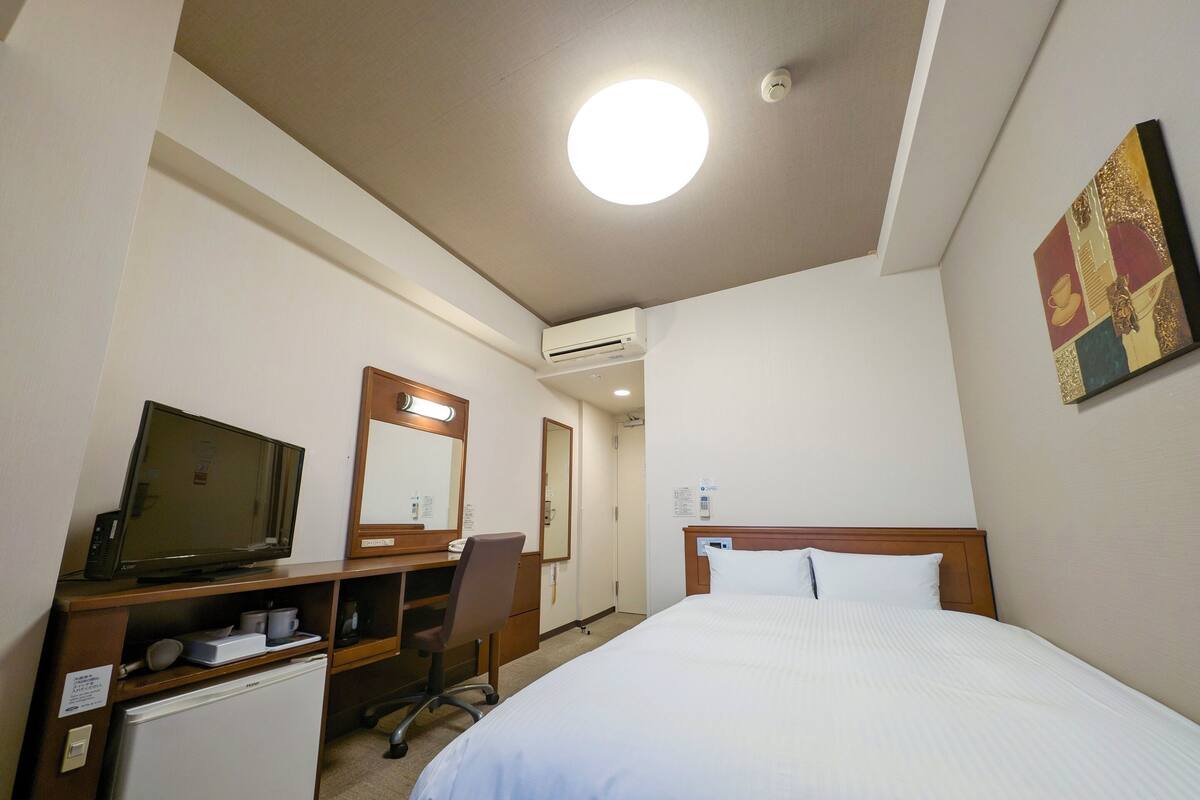 Ett hotellrum med en säng, ett skrivbord med en tv, en stol, ett litet kylskåp och en spegel.