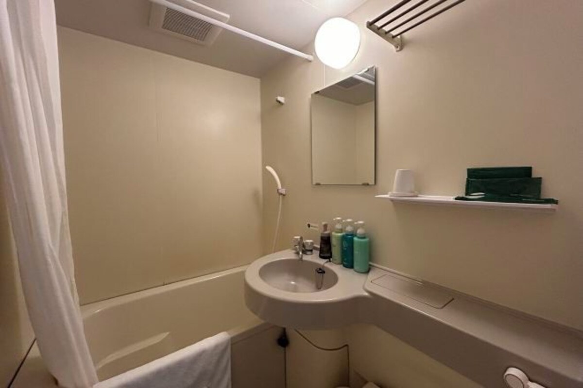 Une salle de bain avec un lavabo, un miroir et une douche. On trouve des articles de toilette sur le lavabo et une serviette est accrochée au mur.